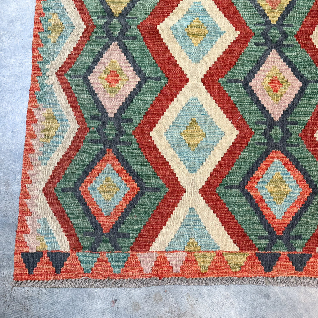 A close-up of a woven kilim rug showing its geometric diamond pattern in red, green, cream, light blue, and orange, with a fringed edge on a concrete floor.