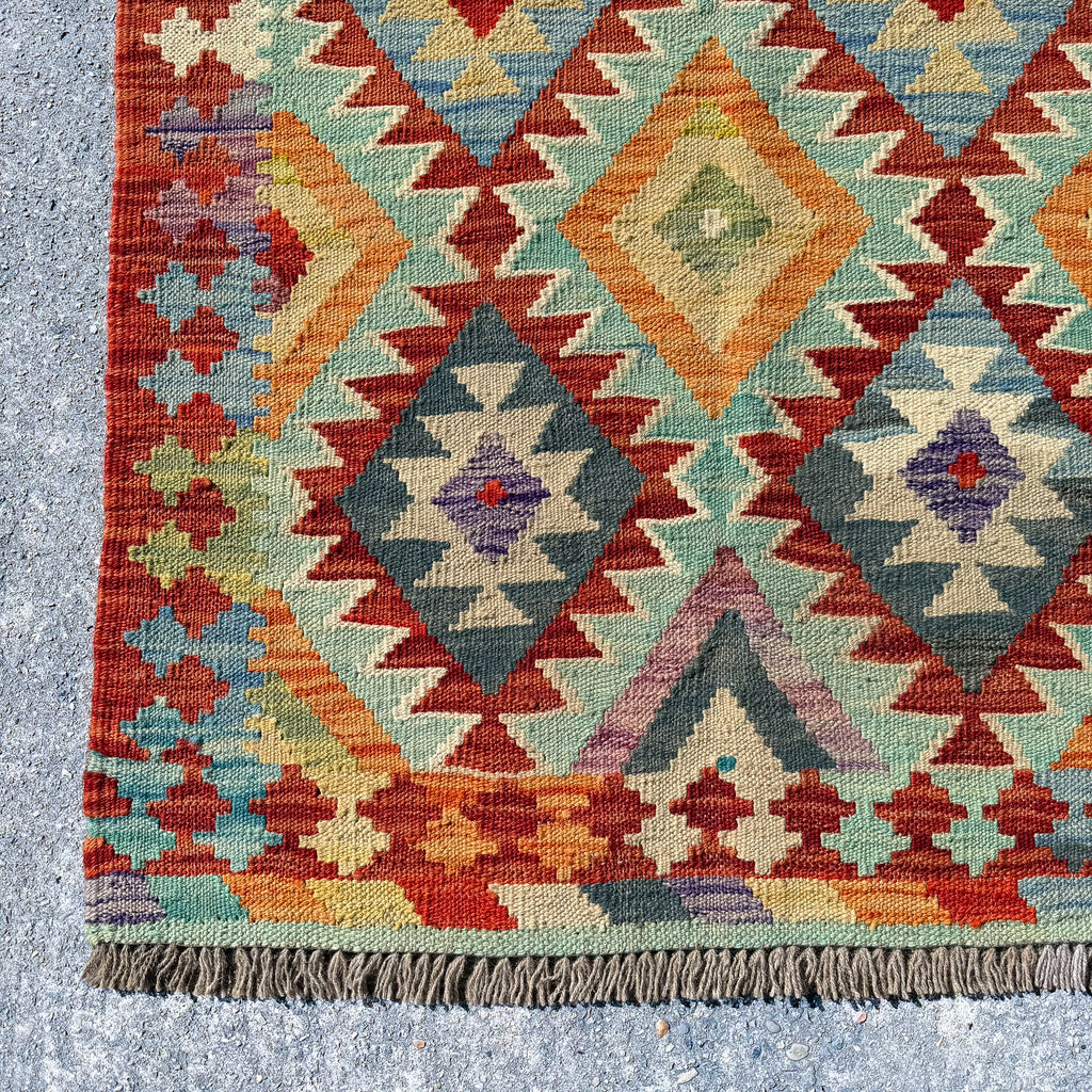 A close-up of a kilim rug showing its detailed geometric pattern and fringed edge, laid on a gray concrete floor.