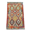 A rectangular kilim rug with a geometric diamond pattern in red, orange, beige, and blue, laid flat on a textured gray concrete surface.