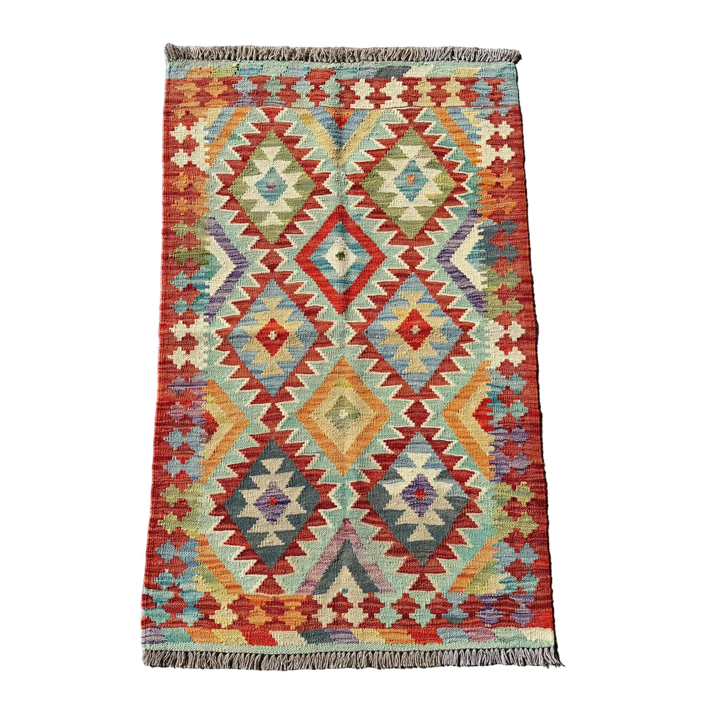 A rectangular kilim rug with a geometric diamond pattern in red, orange, beige, and blue, laid flat on a textured gray concrete surface.