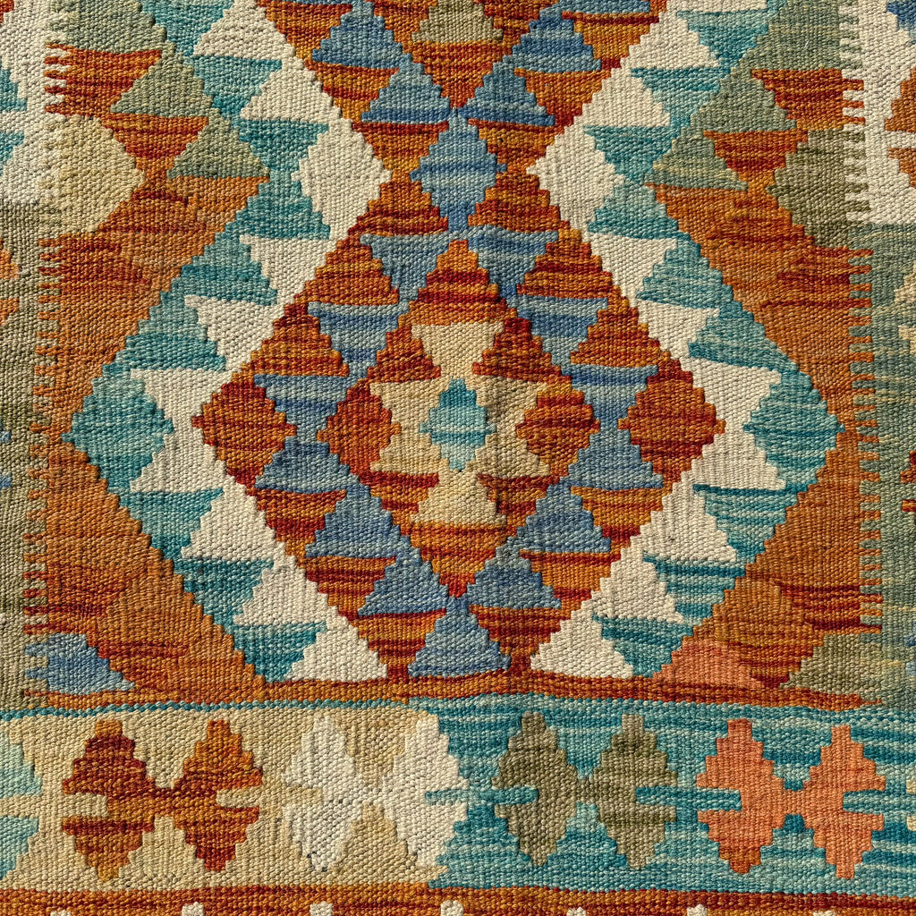 A close-up of a woven textile with a geometric diamond pattern in rust orange, teal blue, cream, and muted olive green.
