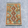 A rectangular kilim-style rug with a geometric diamond pattern in terracotta orange, teal blue, beige, and cream, shown flat on a textured gray concrete surface.
