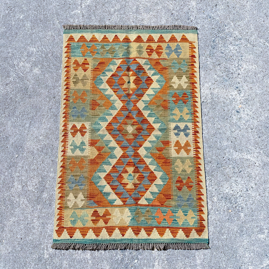 A rectangular kilim-style rug with a geometric diamond pattern in terracotta orange, teal blue, beige, and cream, shown flat on a textured gray concrete surface.