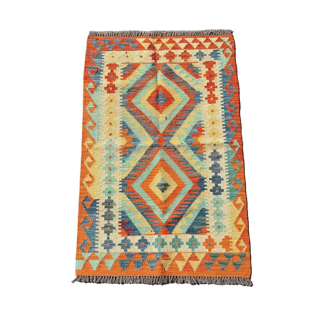 A rectangular vintage kilim rug with a geometric diamond pattern in orange, beige, teal, and blue, laid flat on a gray concrete surface.