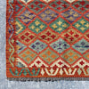 A close-up of a vintage kilim rug with a geometric diamond pattern in terracotta red, teal green, beige, orange, and purple, finished with dark gray fringe.