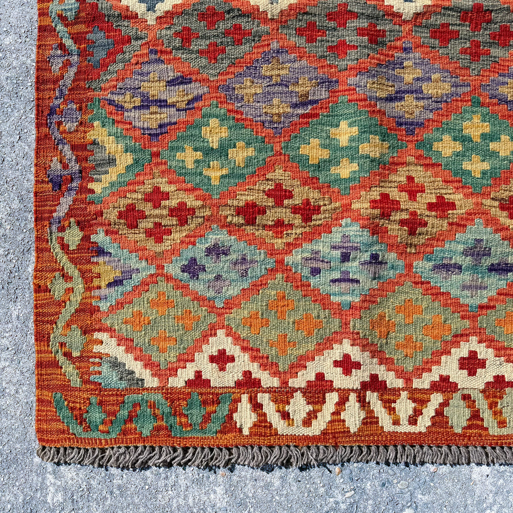 A close-up of a vintage kilim rug with a geometric diamond pattern in terracotta red, teal green, beige, orange, and purple, finished with dark gray fringe.