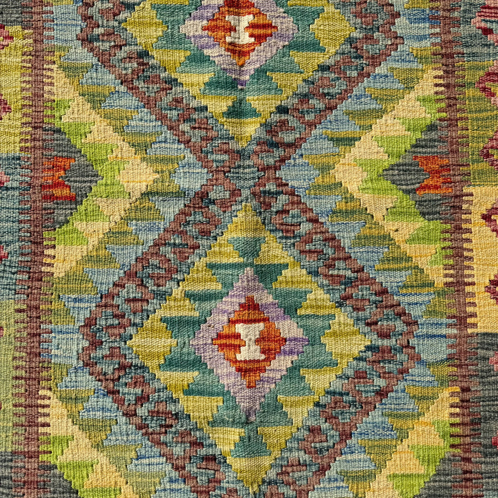 A close-up of a woven geometric rug with a Southwestern-style diamond pattern in muted green, blue, yellow, brown, and red tones.