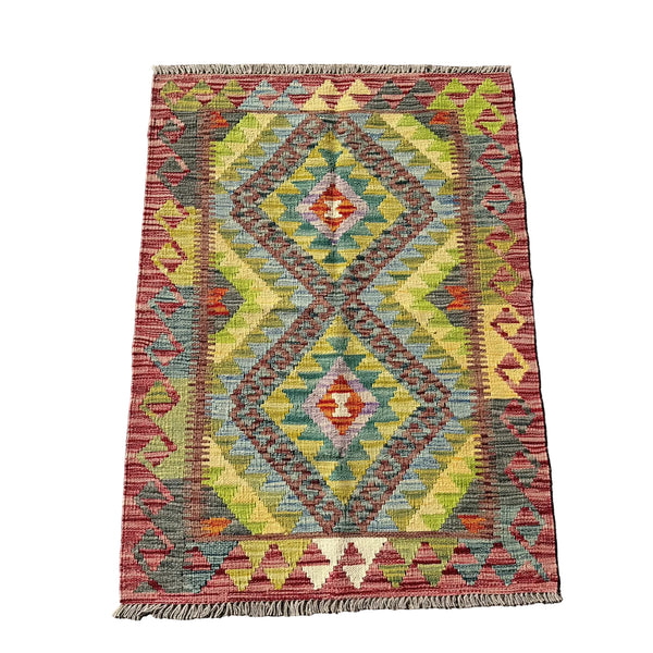 A rectangular kilim rug with a geometric diamond pattern in muted red, yellow, green, blue, and brown, laid flat on a gray concrete surface.