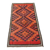 A rectangular woven kilim rug with a bold geometric diamond pattern in red, orange, and black, featuring a decorative border, laid flat on a gray concrete surface.