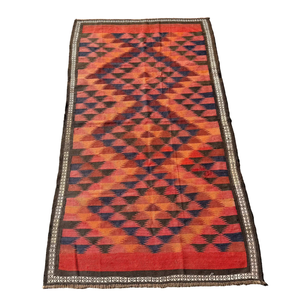 A rectangular woven kilim rug with a bold geometric diamond pattern in red, orange, and black, featuring a decorative border, laid flat on a gray concrete surface.