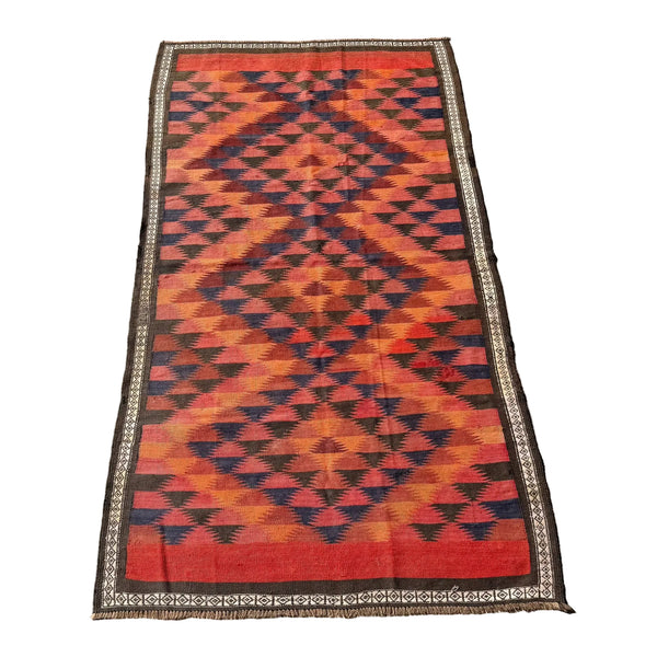 A rectangular woven kilim rug with a bold geometric diamond pattern in red, orange, and black, featuring a decorative border, laid flat on a gray concrete surface.