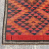 A close-up of a kilim rug with a geometric pattern in orange, red, and dark brown, featuring a decorative border and fringed edge on a gray concrete floor.