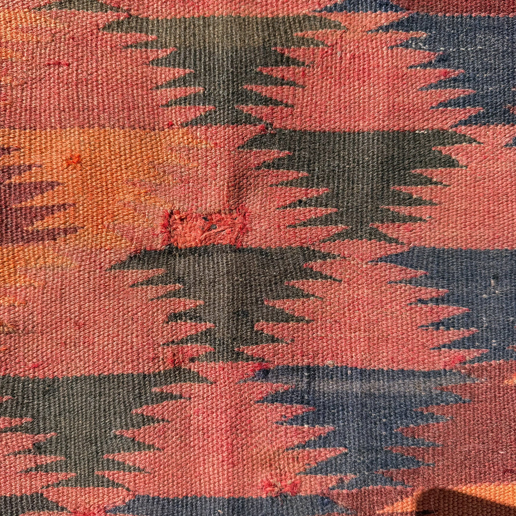 A close-up of a woven textile with a geometric pattern in terracotta pink, orange, dark green, and navy blue.