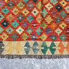 A close-up of a woven kilim rug with a vibrant orange-red background, featuring a multicolor geometric diamond pattern and a fringed border, laid flat on a gray textured surface.