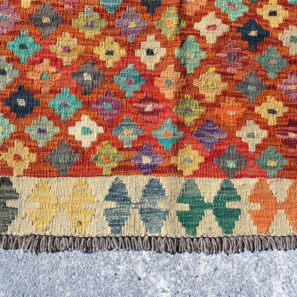 A close-up of a woven kilim rug with a vibrant orange-red background, featuring a multicolor geometric diamond pattern and a fringed border, laid flat on a gray textured surface.