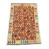 A rectangular kilim rug with a geometric diamond pattern in orange, red, teal, green, and yellow, laid flat on a textured gray concrete surface.