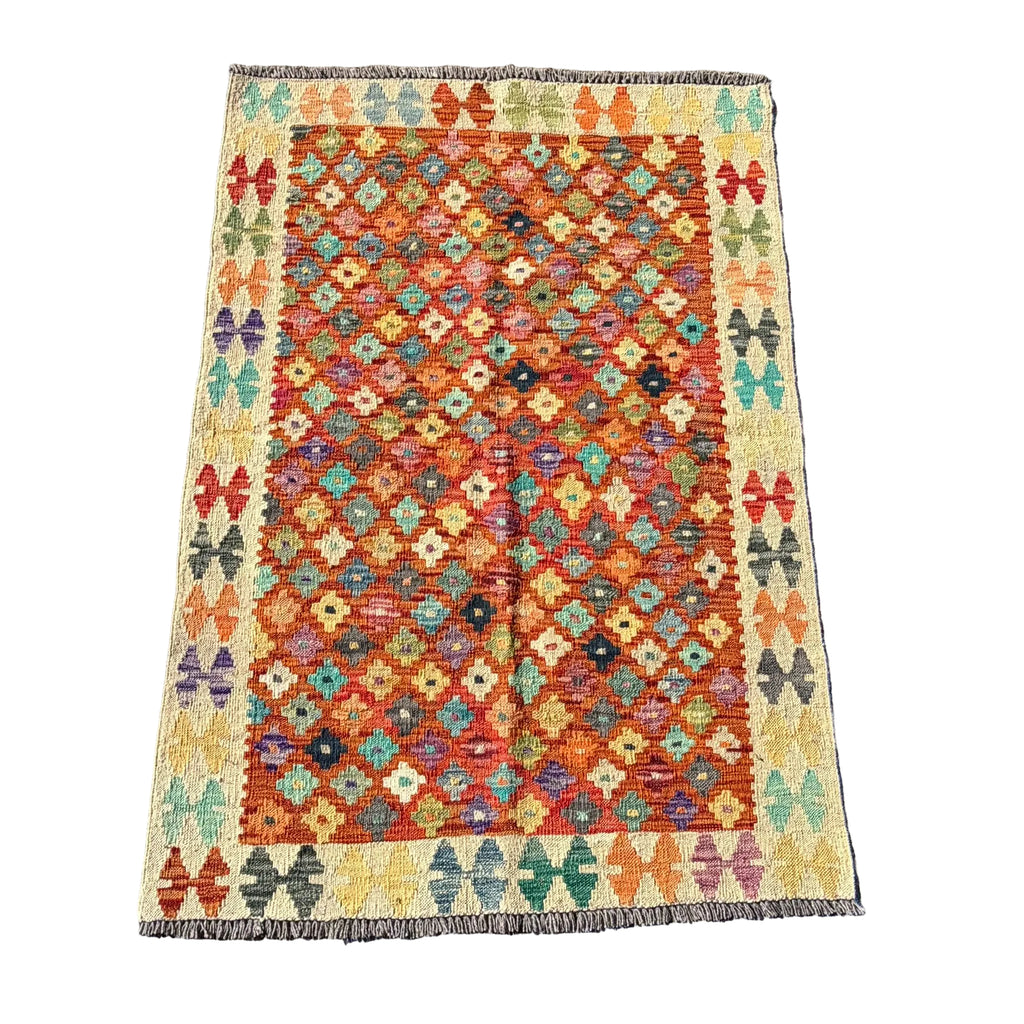 A rectangular kilim rug with a geometric diamond pattern in orange, red, teal, green, and yellow, laid flat on a textured gray concrete surface.