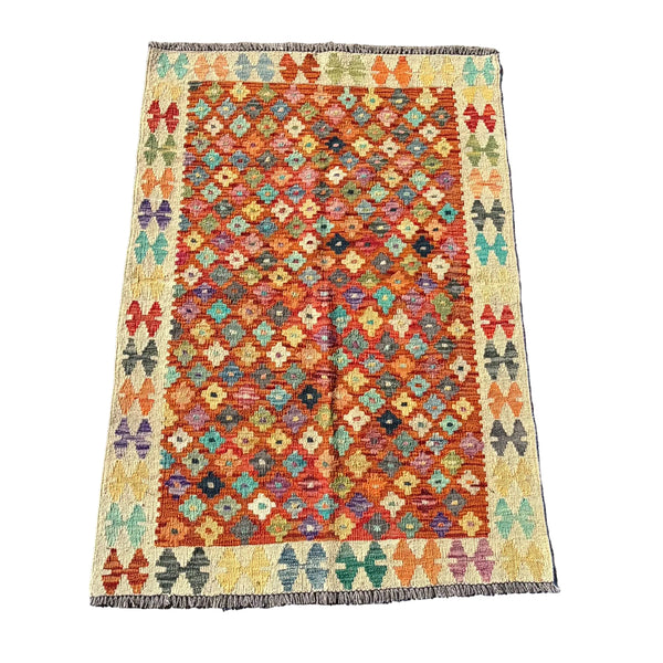 A rectangular kilim rug with a geometric diamond pattern in orange, red, teal, green, and yellow, laid flat on a textured gray concrete surface.