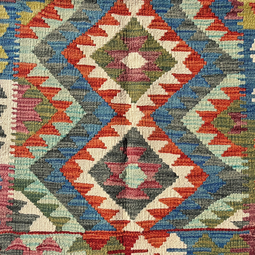 A close-up of a woven kilim textile with a geometric diamond pattern in red, blue, gray, green, pink, and cream.