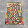 A rectangular kilim rug with a colorful geometric diamond pattern, laid flat on a gray concrete surface.