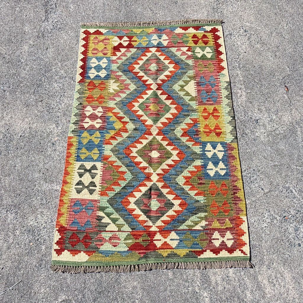 A rectangular kilim rug with a colorful geometric diamond pattern, laid flat on a gray concrete surface.