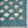 A close-up of a woven rug or textile with a teal-blue base, repeating diamond motifs in cream, beige, and tan, and a fringed edge visible at the top.