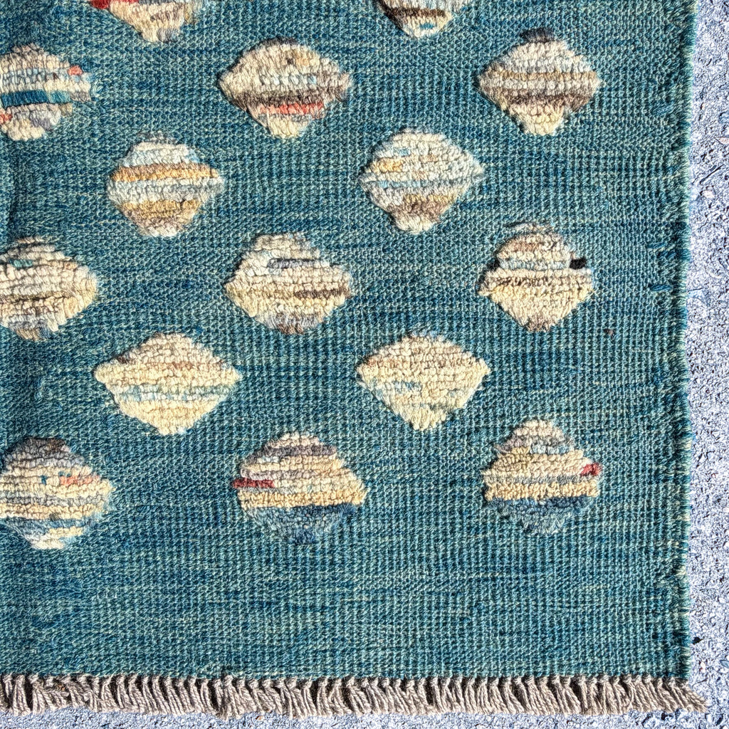 A close-up of a woven rug or textile with a teal-blue base, repeating diamond motifs in cream, beige, and tan, and a fringed edge visible at the top.