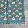 A close-up of a woven rug corner showing a teal blue field with repeating diamond motifs in cream, rust, orange, and brown, finished with a fringed edge on a gray stone surface.