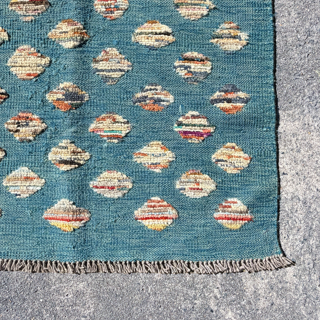 A close-up of a woven rug corner showing a teal blue field with repeating diamond motifs in cream, rust, orange, and brown, finished with a fringed edge on a gray stone surface.