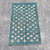 A rectangular blue kilim rug with a repeating pattern of cream and beige diamond motifs, laid flat on a textured gray concrete surface.