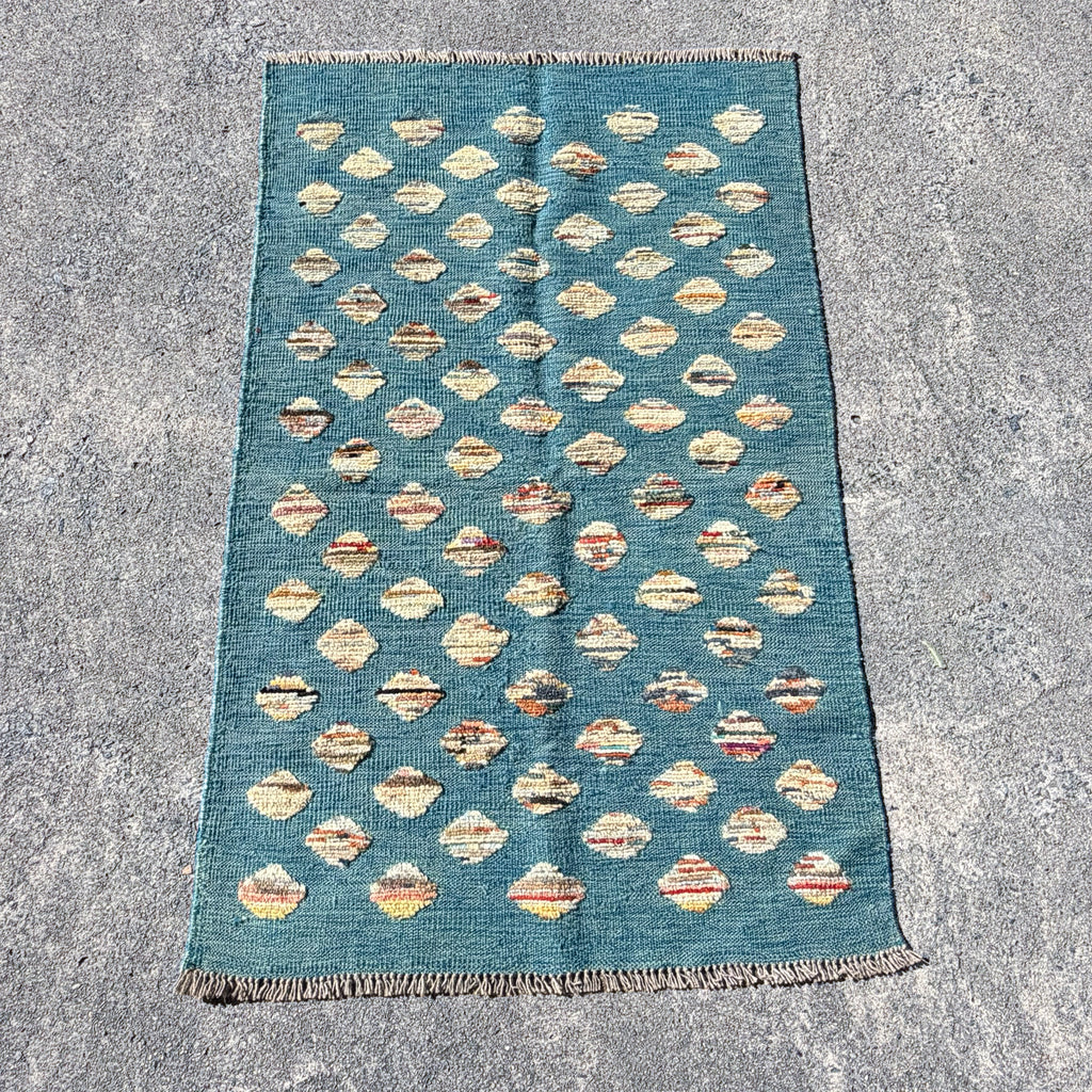 A rectangular blue kilim rug with a repeating pattern of cream and beige diamond motifs, laid flat on a textured gray concrete surface.