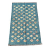 A rectangular blue kilim rug with a repeating pattern of cream and beige diamond motifs, laid flat on a textured gray concrete surface.
