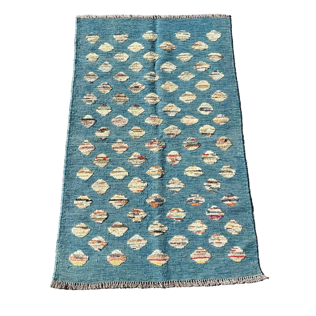 A rectangular blue kilim rug with a repeating pattern of cream and beige diamond motifs, laid flat on a textured gray concrete surface.