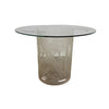 A round glass-top side table with a frosted, etched glass pedestal base featuring a botanical leaf pattern, set against a plain white wall and concrete floor.