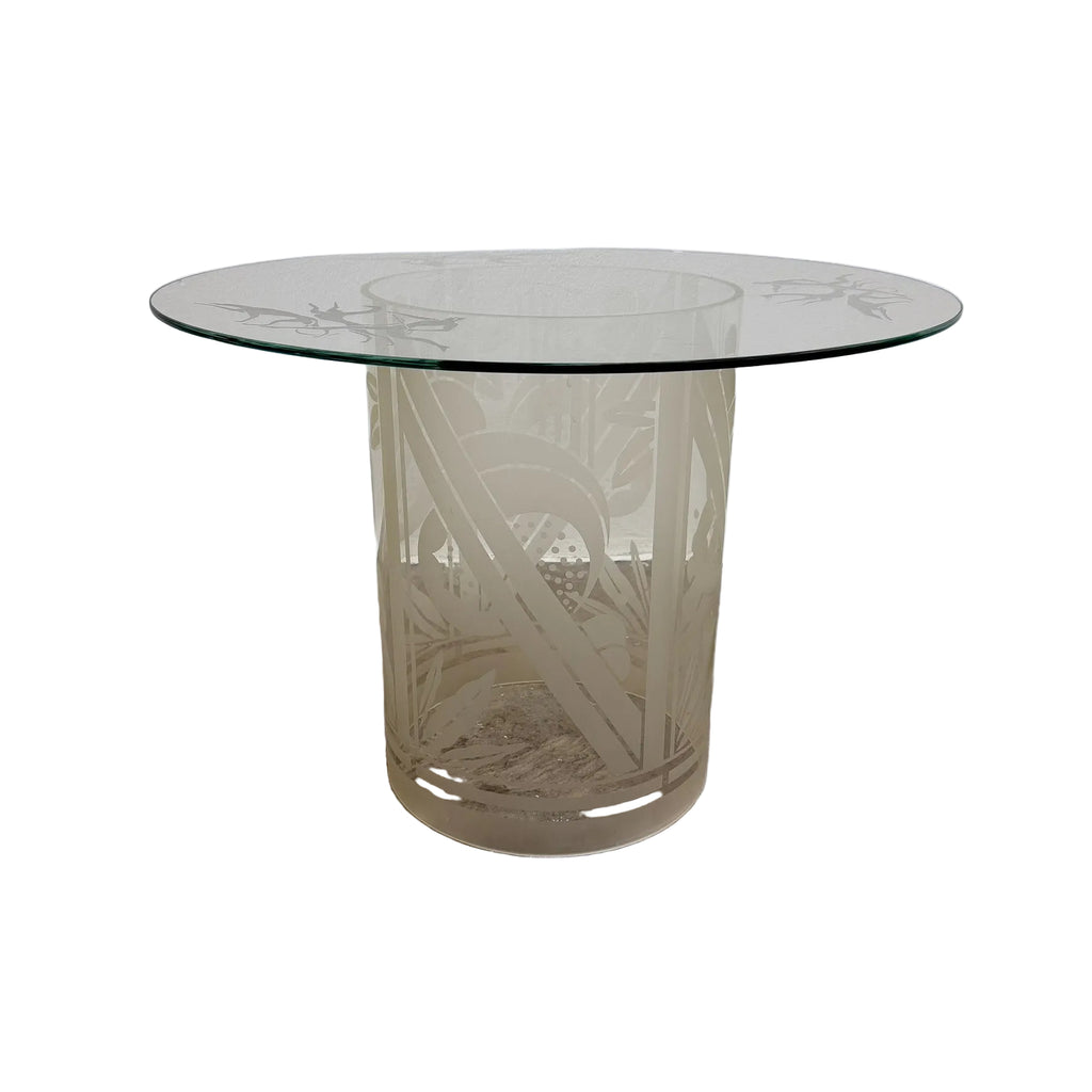 A round glass-top side table with a frosted, etched glass pedestal base featuring a botanical leaf pattern, set against a plain white wall and concrete floor.