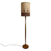 A vintage mid-century modern floor lamp with a tall, slender wooden stand and a cylindrical drum shade. The shade is beige with a dark brown geometric pattern near the bottom, and the lamp base and stand are made of medium brown wood.