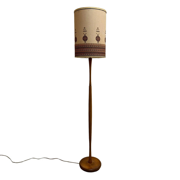 A vintage mid-century modern floor lamp with a tall, slender wooden stand and a cylindrical drum shade. The shade is beige with a dark brown geometric pattern near the bottom, and the lamp base and stand are made of medium brown wood.