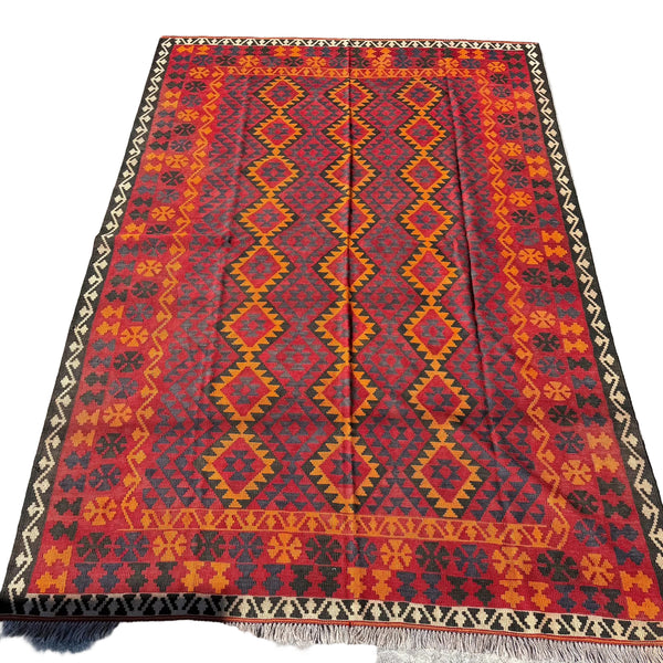 A rectangular kilim area rug with a bold geometric pattern in red, orange, yellow, and black, laid flat on a gray concrete surface.