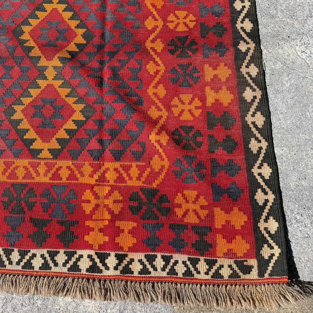 A close-up of a woven kilim rug corner showing a red field with black and ochre geometric diamond patterns, a dark border, and beige fringe on a gray concrete surface.