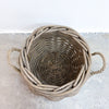 A round woven basket made from natural driftwood or weathered wood, featuring two twisted side handles and a rustic, weathered gray-brown finish, photographed from a top-down angle on a concrete floor against a white wall.