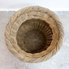 A round woven basket made of natural light brown wicker or rattan, photographed from directly above on a light gray concrete floor.