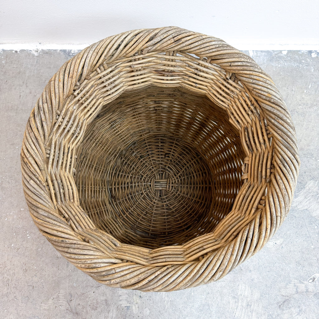 A round woven basket made of natural light brown wicker or rattan, photographed from directly above on a light gray concrete floor.