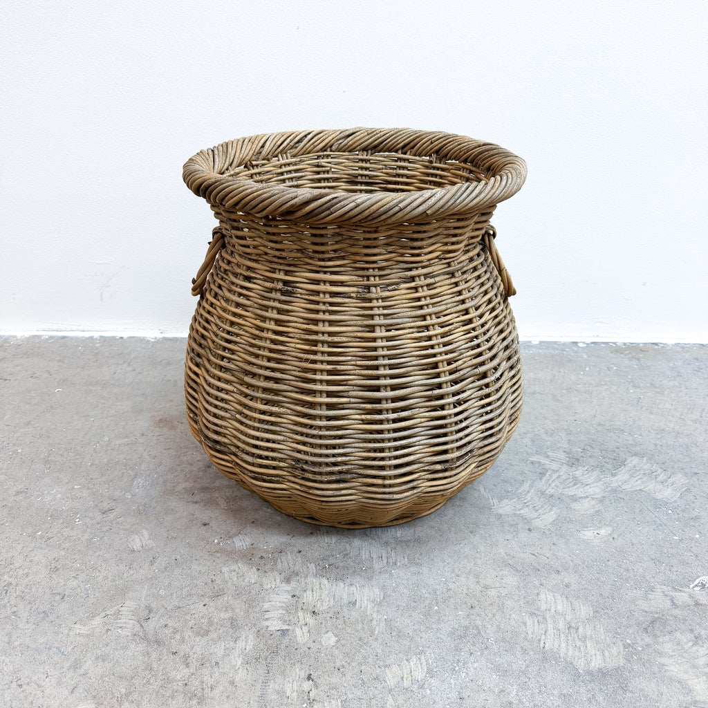 A round woven wicker basket with a twisted rope rim and side handles, standing on a gray concrete floor against a white wall.