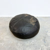 A round black leather pouf ottoman with visible wear, scuffs, and dirt marks, sitting on a concrete floor against a white wall.