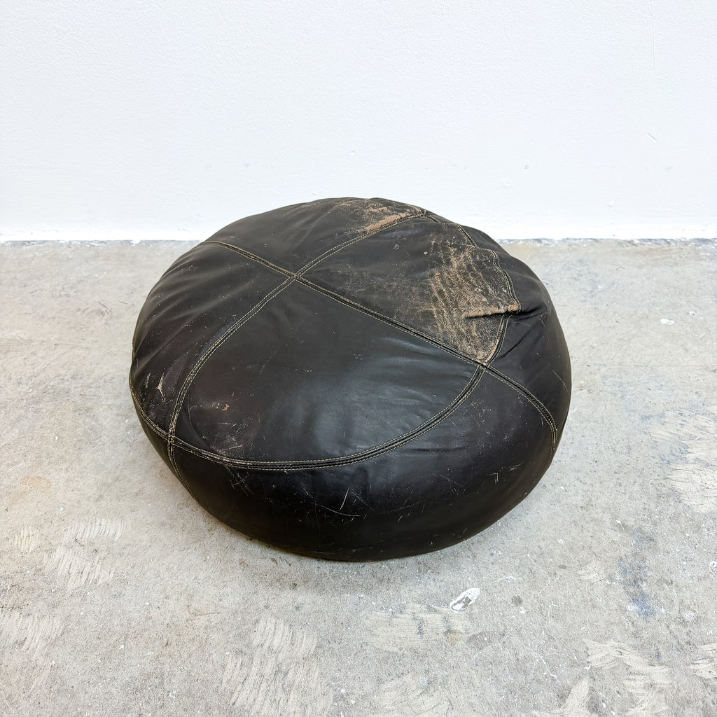 A round black leather pouf ottoman with visible wear, scuffs, and dirt marks, sitting on a concrete floor against a white wall.