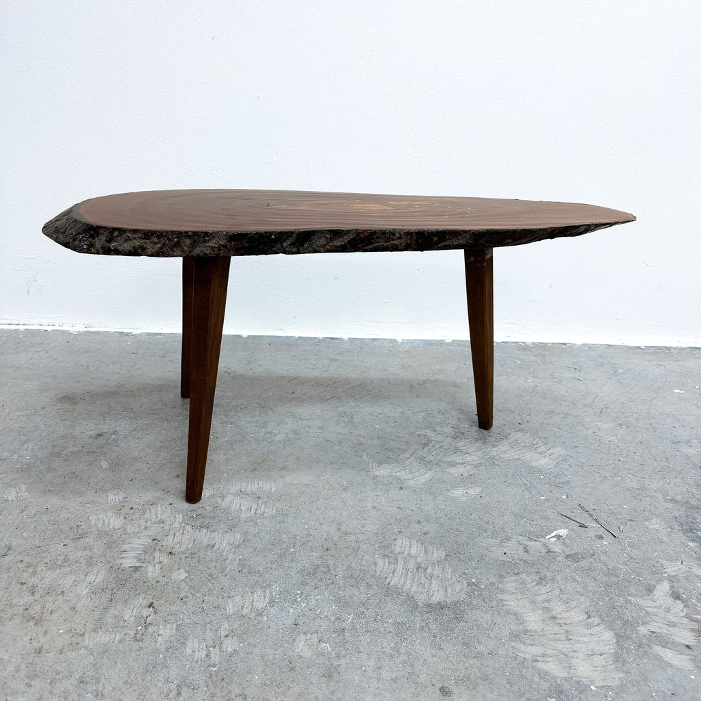 A mid-century modern live-edge wooden coffee table with a dark brown top and legs, set against a plain white wall and concrete floor.