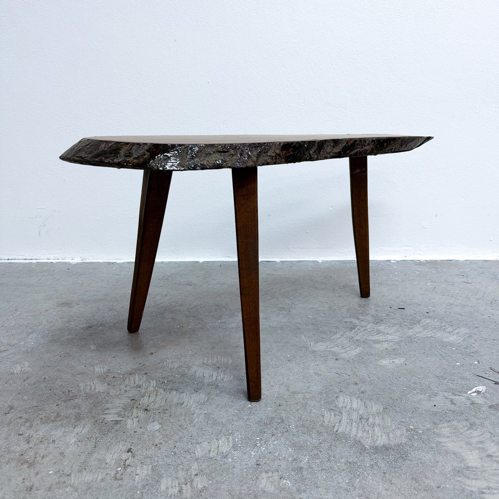 A mid-century modern coffee table with a dark, irregularly shaped live-edge wood top and three tapered wooden legs, set against a plain white wall and concrete floor.