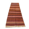A long rectangular striped wool runner rug laid flat on a gray concrete surface outdoors, with grass visible at the right edge. The rug has horizontal bands in rust red, brown, orange, beige, and muted pink, with fringed ends.