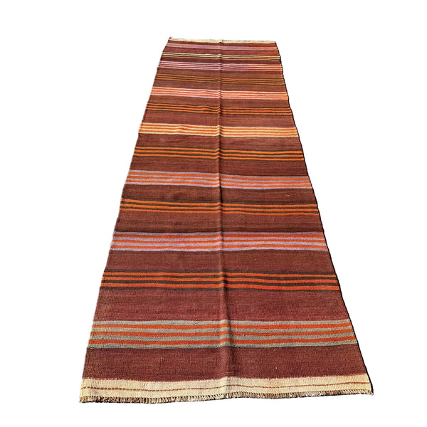 A long rectangular striped wool runner rug laid flat on a gray concrete surface outdoors, with grass visible at the right edge. The rug has horizontal bands in rust red, brown, orange, beige, and muted pink, with fringed ends.