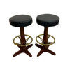 Two vintage-style bar stools with round black upholstered seats, dark wood pedestal bases, and brass footrest rings, shown on a concrete floor against a white wall.
